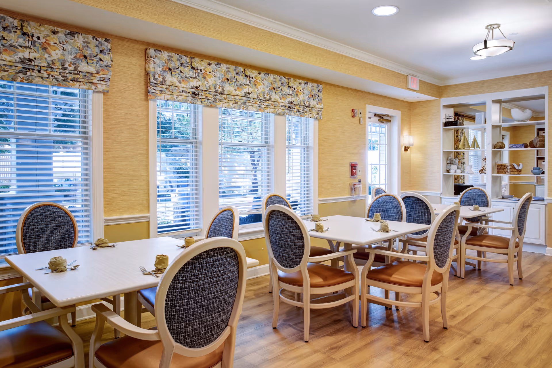 A bright dining room with multiple tables and chairs arranged neatly. Each table has napkins and cutlery set for dining. The room features large windows with floral-patterned valances, light yellow walls, wooden flooring, and a white shelving unit with decorative items.
