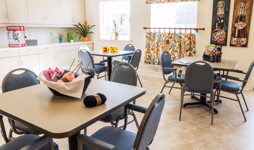 Bright communal dining/activity room with several tables and chairs, yarn and craft supplies on a table, and cabinets along the wall.