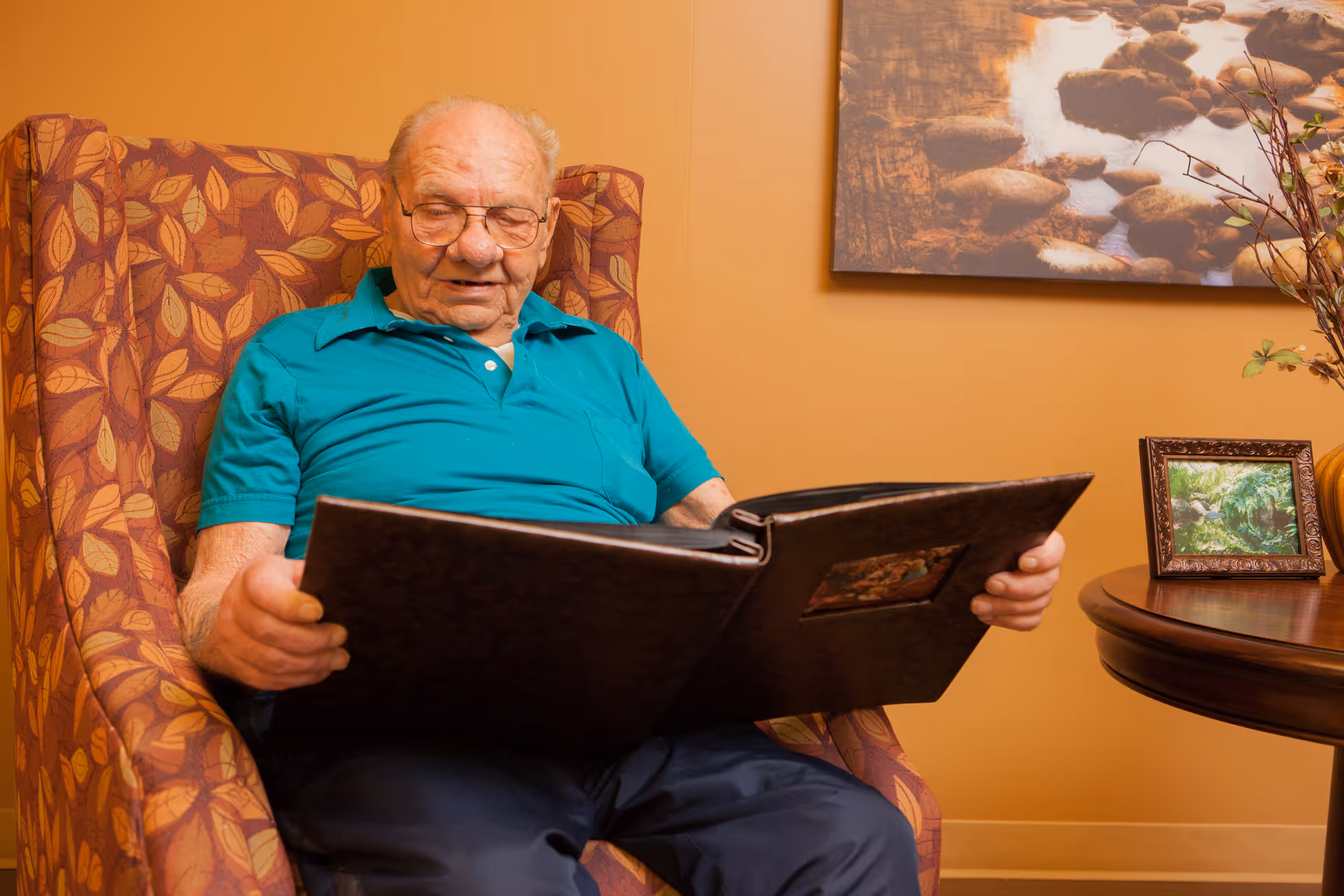 An elderly man wearing glasses and a teal polo shirt sits in a patterned armchair, looking at a large photo album. Behind him is a wall with a framed picture of a rocky stream and a small round wooden table with a framed photo and decorative branches.
