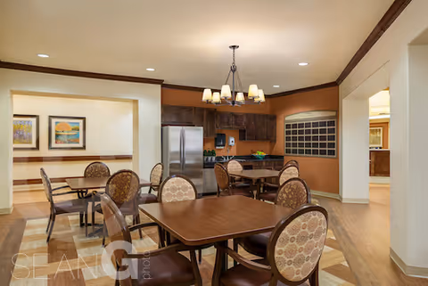 Interior view of a dining area in a senior living facility with multiple wooden tables and cushioned chairs arranged neatly. The room features warm lighting with a chandelier, wood cabinetry, a stainless steel refrigerator, and framed artwork on the walls. The flooring is wood, and the space appears clean and inviting.