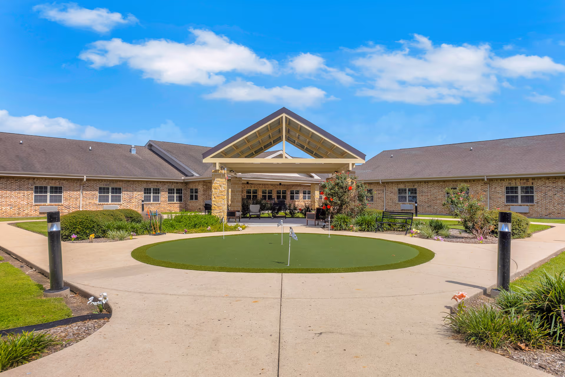 Courtyard and covered entrance of a single-story brick building with a circular putting green, walkways, benches, and landscaping.