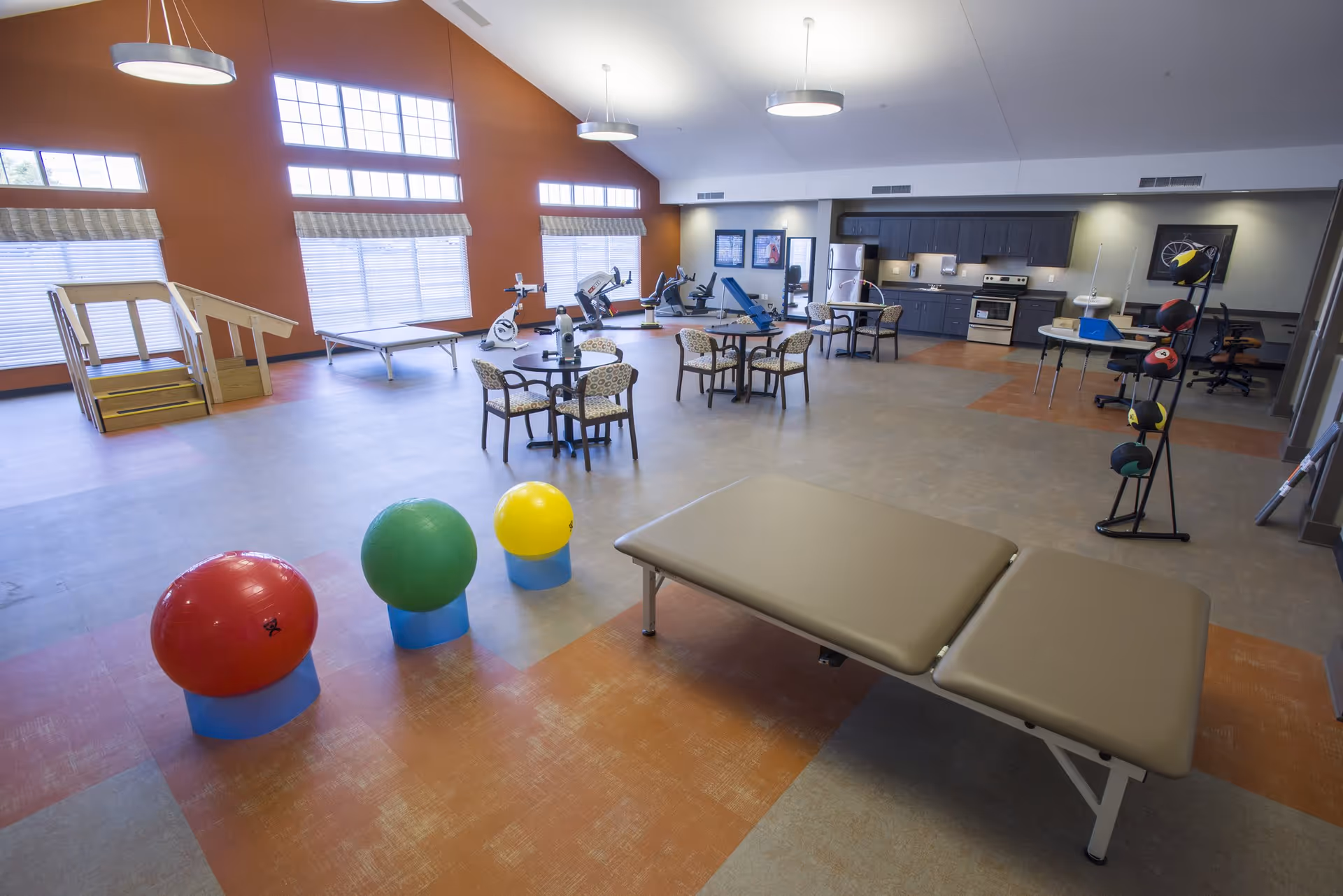 A spacious rehabilitation or physical therapy room with exercise balls, therapy tables, stationary bikes, and other exercise equipment. There are several round tables with chairs, a small wooden staircase for therapy, and a kitchenette area with cabinets, a stove, and a refrigerator. Large windows with blinds allow natural light to fill the room.