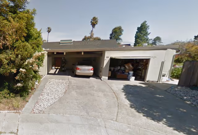 Exterior view of a single-story residential building with a two-car garage, one side open showing a parked car and the other side filled with various stored items. The driveway is concrete with a small landscaped area containing white rocks and plants. Trees and shrubs surround the property under a clear sky.