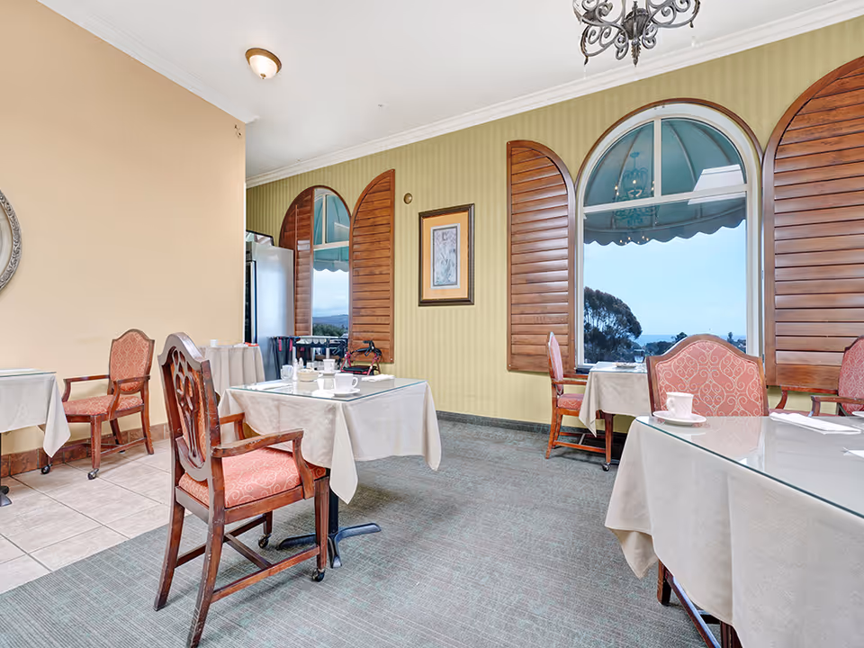 Dining room with tables covered in white tablecloths and wooden chairs with red patterned cushions. Large arched windows with wooden shutters offer a view of trees and a distant landscape under a blue sky. The room has a chandelier, framed artwork on the wall, and a mix of carpet and tiled flooring.