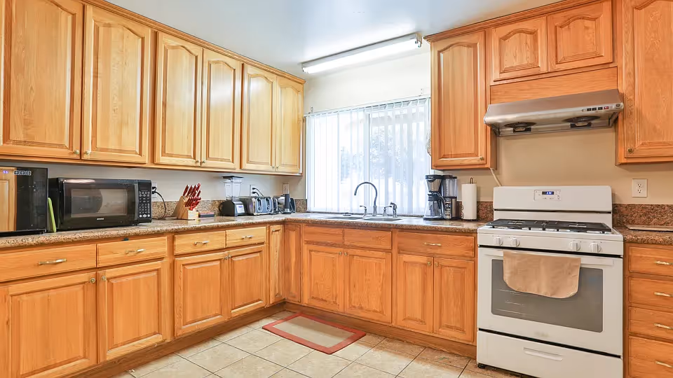 A bright kitchen with wooden cabinets, granite countertops, a white gas stove with a beige towel hanging on the handle, a stainless steel range hood, a coffee maker, a knife block, a microwave, a toaster, a blender, and a window with vertical blinds letting in natural light.