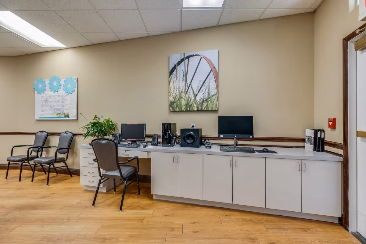 Community room interior with a long cabinet countertop holding two computer monitors, chairs, a plant, and wall art on a beige wall.