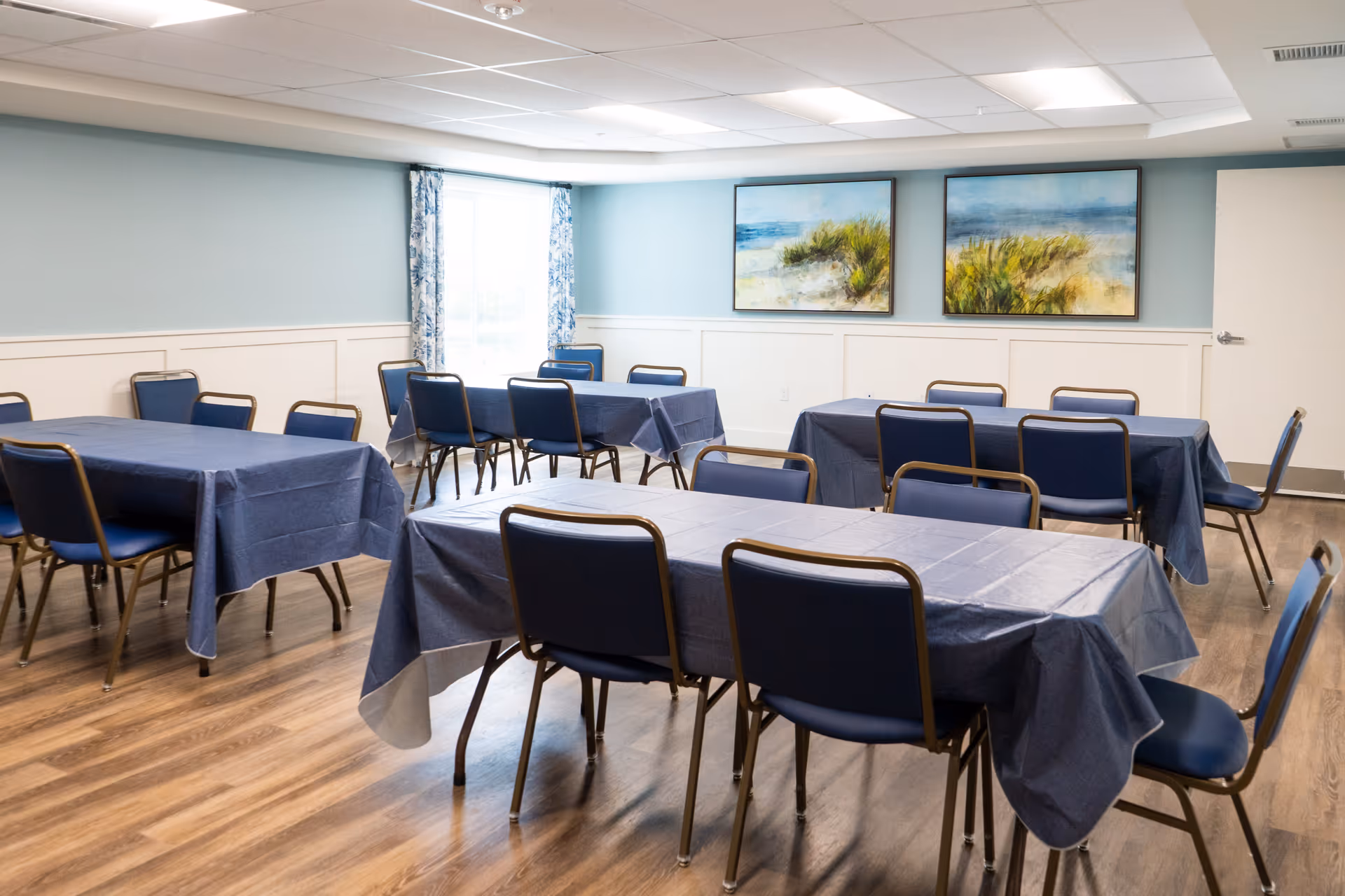 A bright dining/activity room with several tables covered in blue tablecloths and matching chairs, coastal paintings on the wall and a window with patterned curtains.