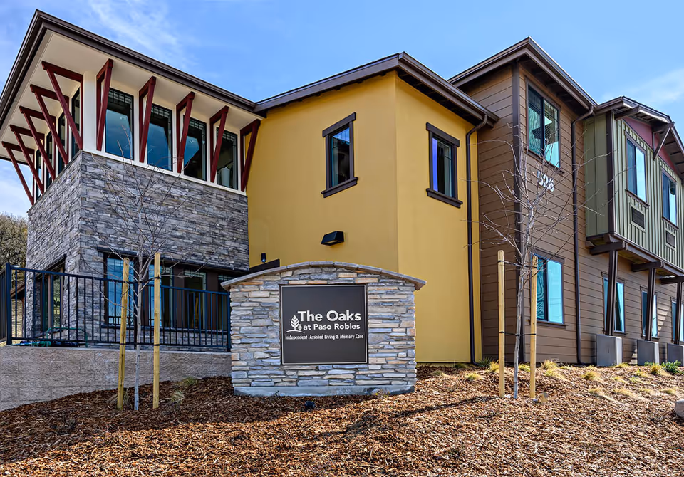 Exterior view of The Oaks at Paso Robles senior living facility showing a modern building with stone and wood siding, multiple windows, and a sign in front that reads 'The Oaks at Paso Robles Independent Assisted Living & Memory Care'.