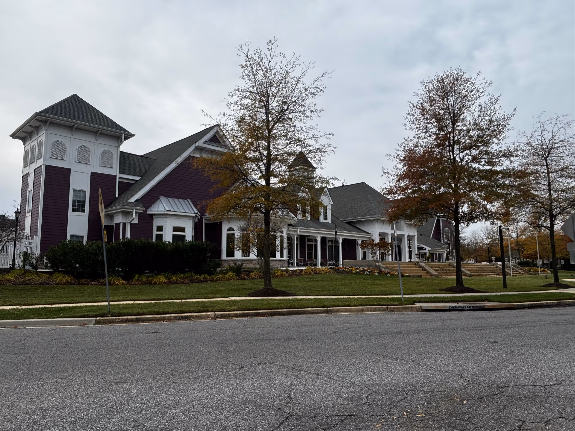 Large purple-and-white senior living building with a covered porch, lawn, and trees along a street under a cloudy sky.