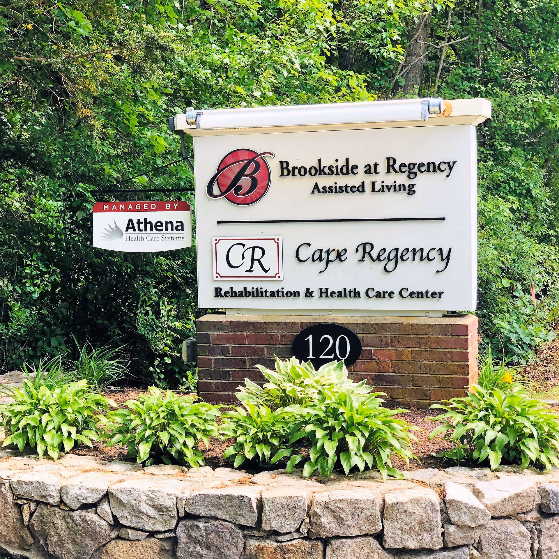 Outdoor sign for Brookside at Regency Assisted Living and Cape Regency Rehabilitation & Health Care Center, surrounded by greenery and plants with a stone wall in front.