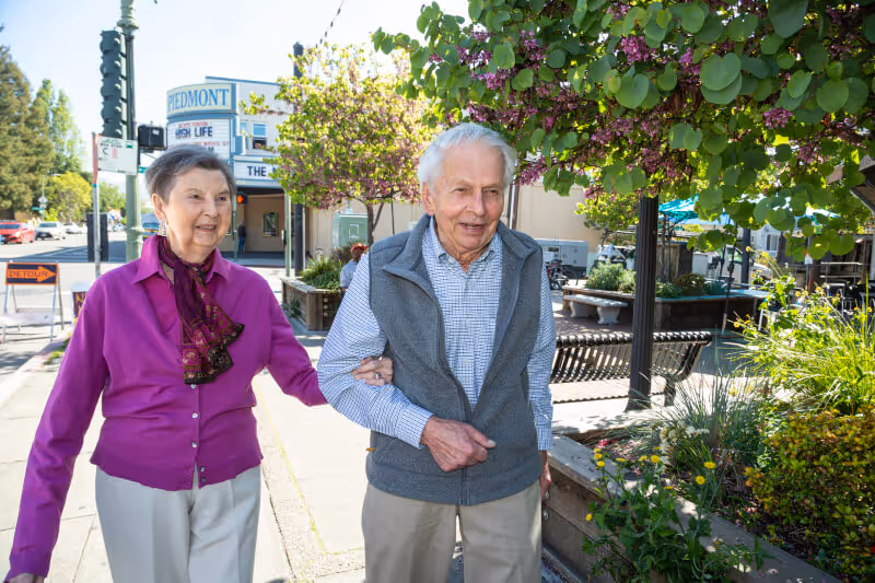 An elderly couple walking arm in arm on a sunny sidewalk next to a garden area with blooming trees and plants. The man is wearing a gray vest over a checkered shirt and beige pants, and the woman is wearing a magenta cardigan with a patterned scarf and light-colored pants. In the background, there is a sign that reads 'Piedmont' and some street signs and buildings.