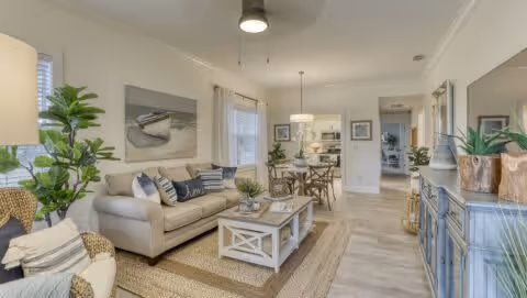 A bright and airy living room with a beige sofa, a wicker chair with cushions, a white coffee table, and a blue sideboard. The room features light-colored walls, large windows with white curtains, indoor plants, and a dining area with a wooden table and chairs in the background.