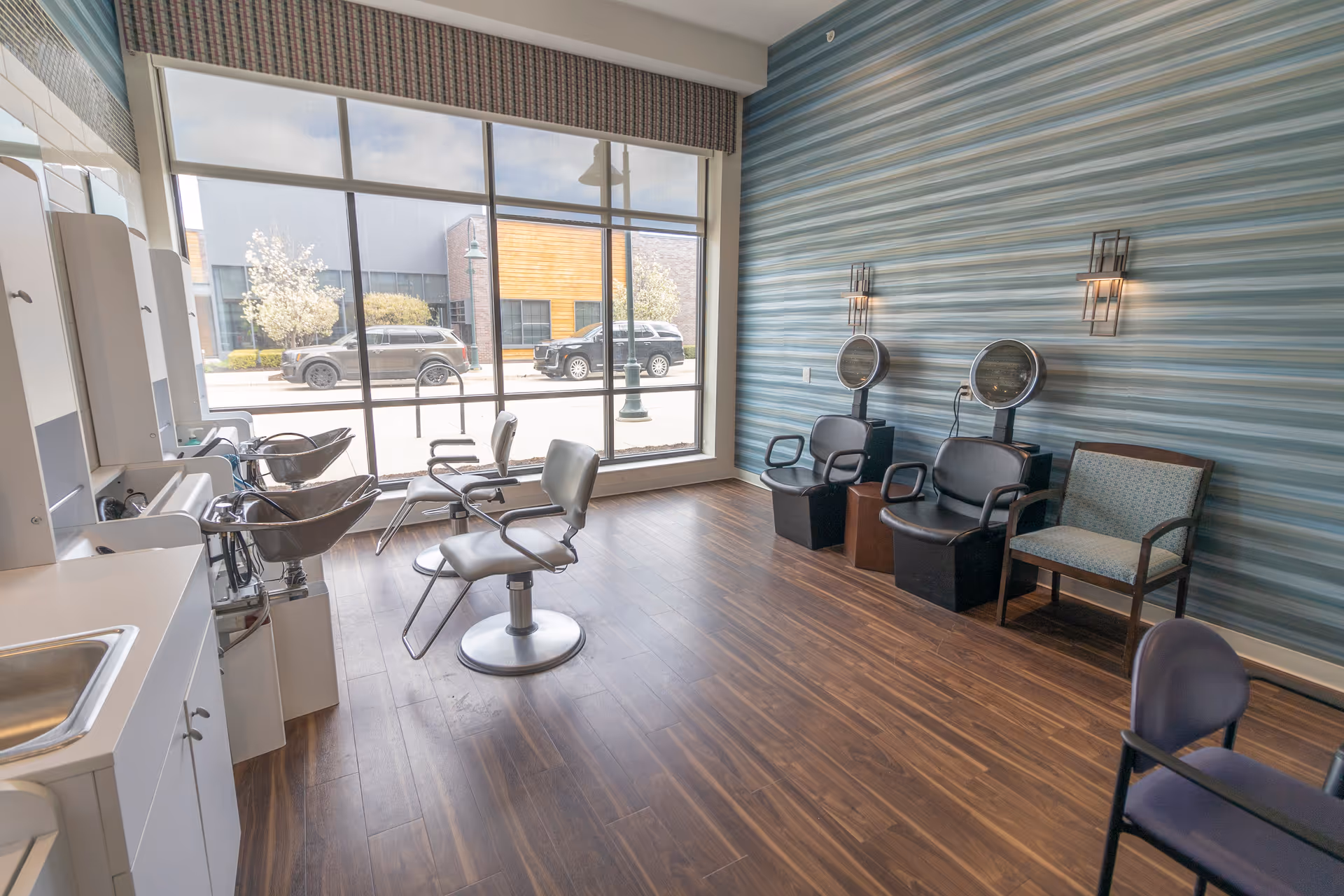 Interior view of a hair salon area with two salon chairs in front of sinks, two hair drying chairs against a blue striped wall, and large windows showing parked cars outside.