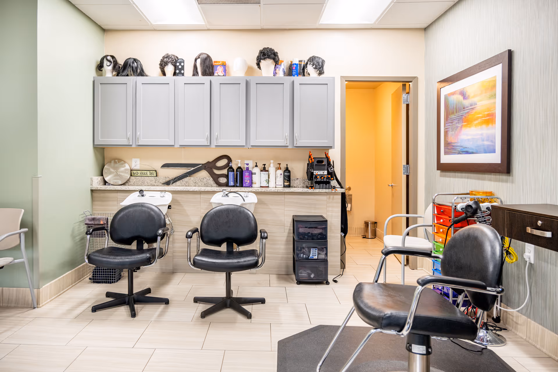 Interior view of a hair salon area with two black salon chairs in front of two hair washing sinks. Above the sinks are gray cabinets with mannequin heads wearing wigs on top. Various hair care products and a large decorative pair of scissors are on the counter. To the right, there is a black salon chair, a colorful drawer organizer, a hair dryer, and a framed painting on the wall. The floor is tiled and there is an open doorway leading to another room.