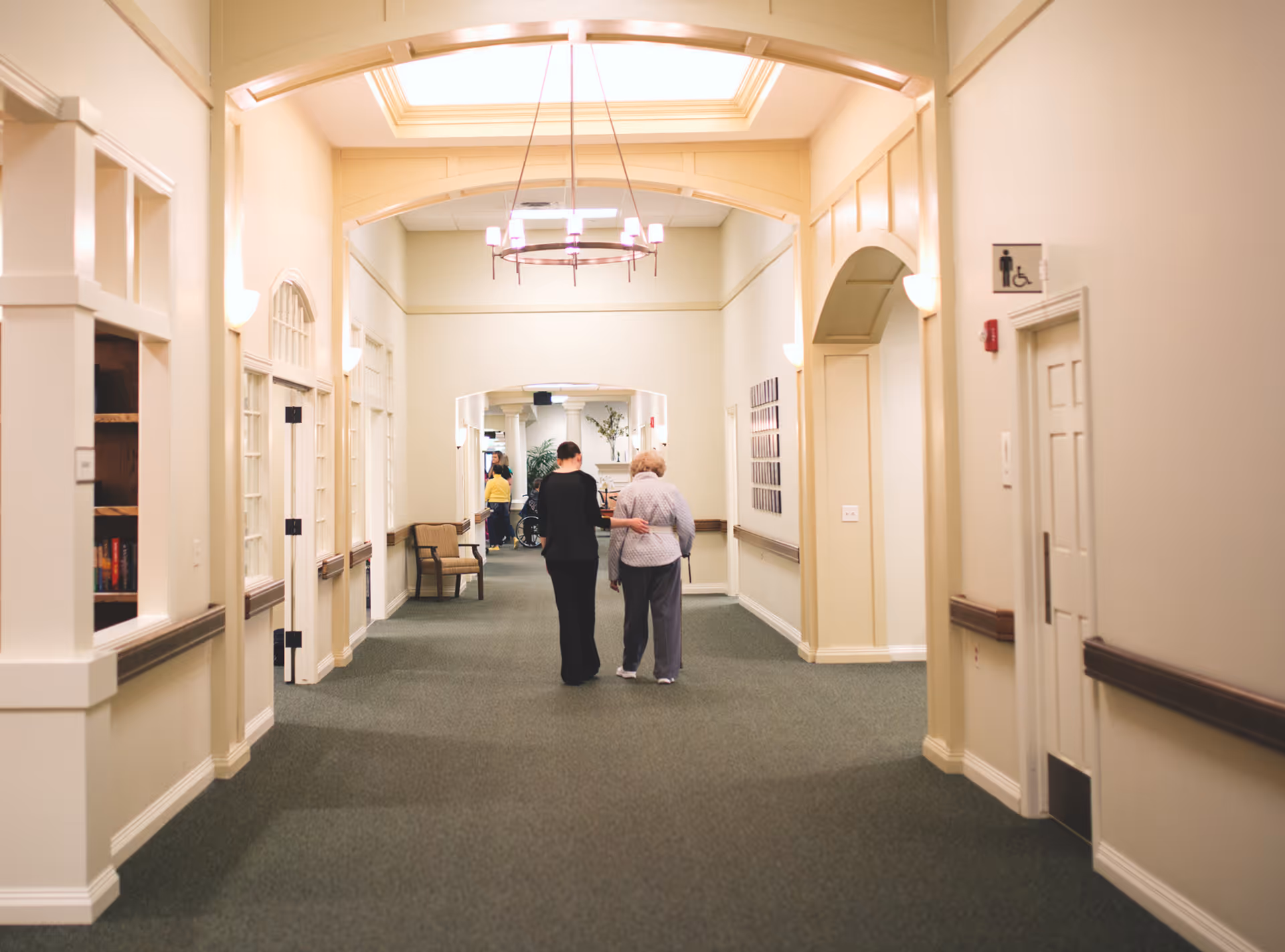 A wide hallway in a senior living facility with beige walls and green carpet. Two people, one elderly woman and a caregiver, walk arm in arm down the hallway. The hallway is well-lit with ceiling lights and wall sconces. There are chairs and framed pictures along the walls, and a restroom sign is visible on the right side.