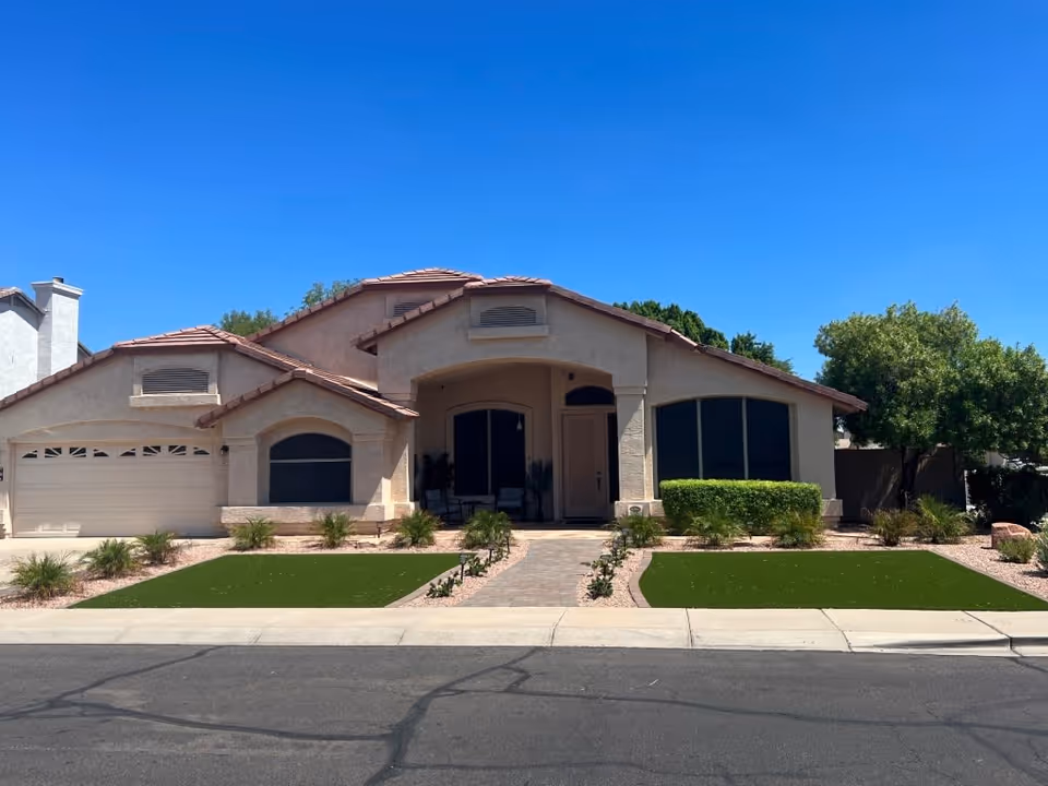 Front exterior view of a single-story house with a tiled roof, two large windows, a garage door on the left, a central entrance with a small porch, and landscaped front yard with green grass patches and shrubs under a clear blue sky.