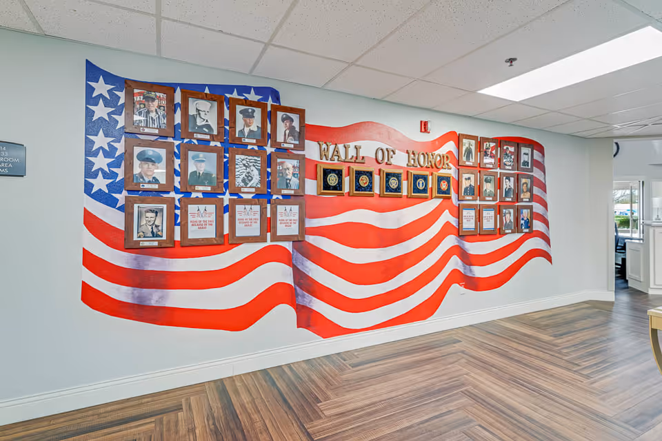 Interior wall of Solstice Senior Living at Bakersfield featuring a large mural of the American flag with framed photos and plaques titled 'Wall of Honor' displaying military personnel and honors.