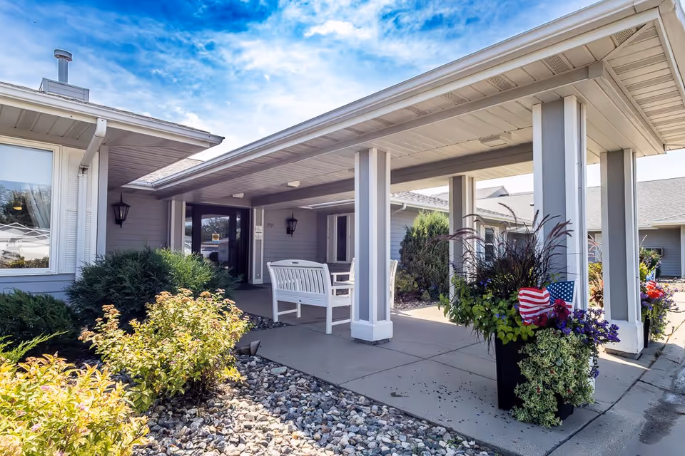 Entrance area of Elison Assisted Living of Minot featuring a covered walkway with white columns, a white bench, and landscaped bushes and flowers including a planter with American flags. The building exterior is light gray with a window and outdoor wall lanterns under a partly cloudy blue sky.