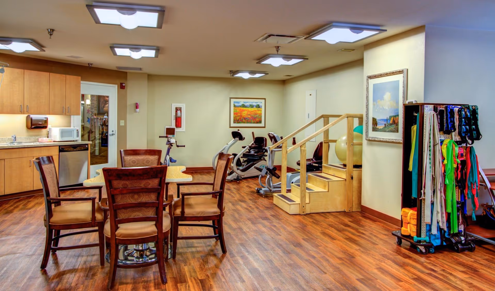 A well-lit room with wooden flooring featuring a small kitchenette with cabinets, a microwave, and a dishwasher on the left. In the center, there is a wooden table surrounded by four cushioned chairs. Towards the back, there are exercise machines and a set of wooden stairs with handrails. On the right side, there is a rack holding various colorful exercise bands and equipment. The walls are decorated with framed landscape paintings.
