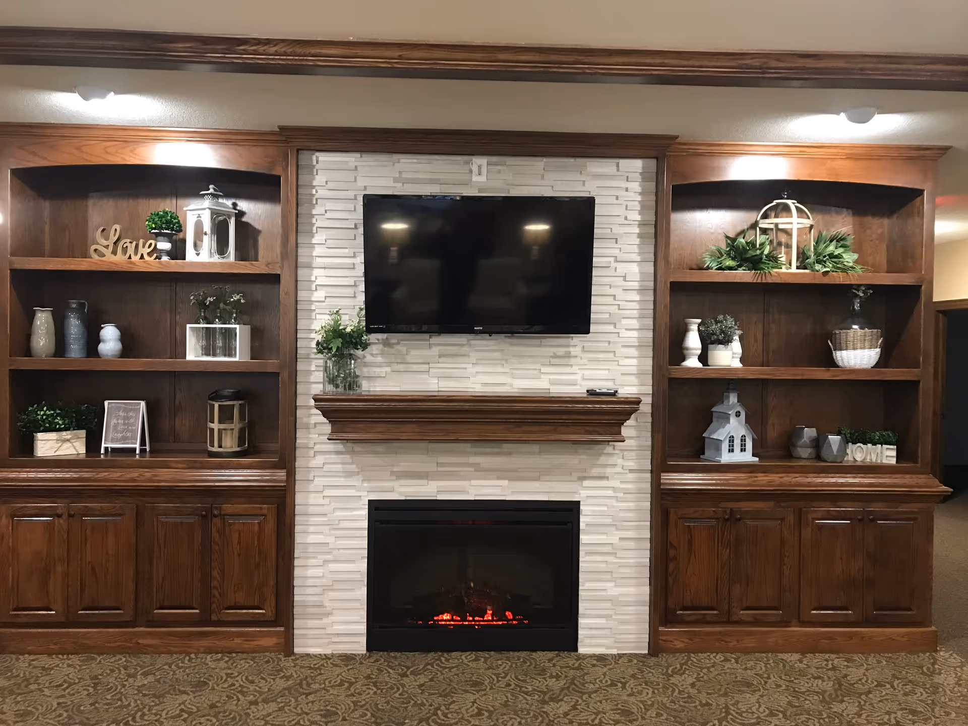 A cozy living room area featuring a wall-mounted flat-screen TV above a wooden mantel and an electric fireplace with glowing embers. On either side of the fireplace are built-in wooden shelves decorated with various plants, vases, lanterns, and decorative signs including the words 'Love' and 'Home'. The floor is carpeted with a patterned design.