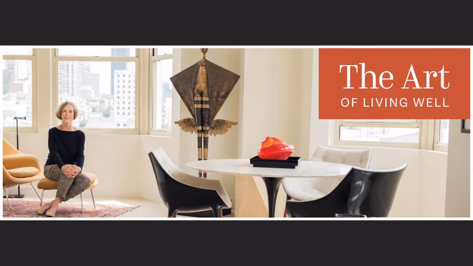 A bright, modern living room with large windows showing a cityscape outside. An elderly woman sits smiling on a mustard-yellow chair on the left side. In the center, there is a round white table with four chairs around it, two black and two white. A red decorative object sits on the table. Behind the table is a tall, abstract wooden sculpture. The top right corner has an orange banner with the text 'The Art of Living Well'.