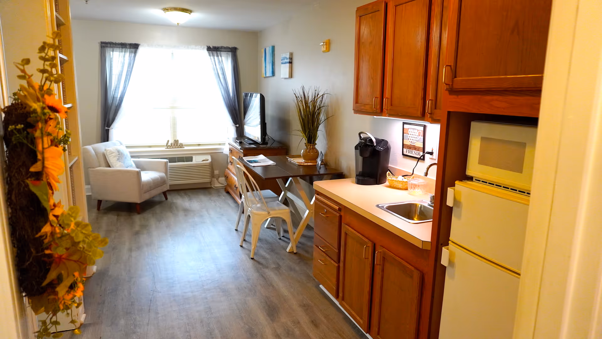 Small studio interior showing a kitchenette on the right, a dining table and TV in the center, and an armchair by a sunlit window at the far end.