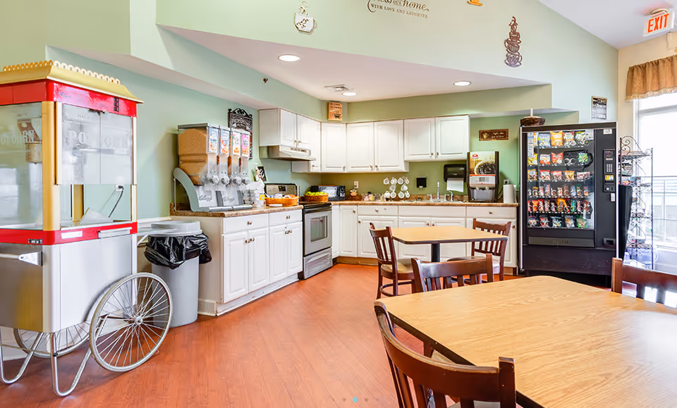 A bright and clean kitchen and dining area in an assisted living facility. The room features white cabinets, a stove, a microwave, and a countertop with cereal dispensers and a coffee machine. There is a popcorn machine on the left side and a vending machine on the right. Several wooden tables and chairs are arranged for dining. The walls are painted light green, and there is an exit door with a window letting in natural light.