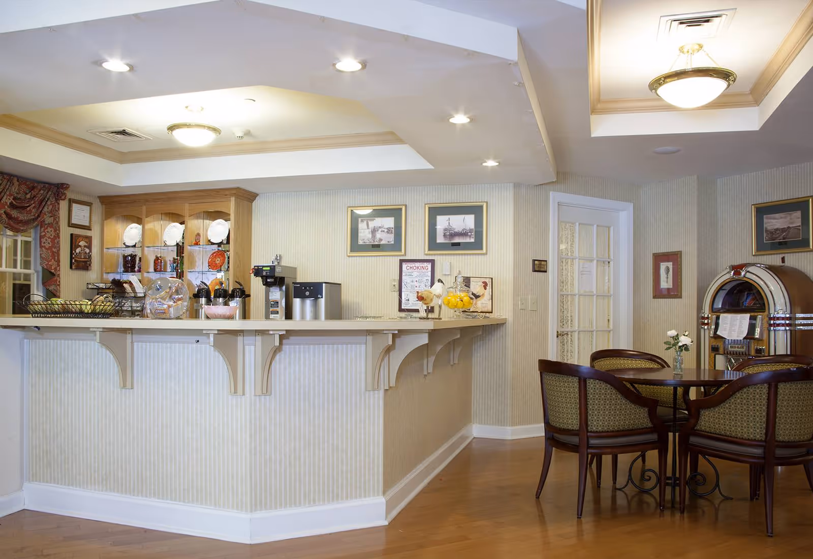 Interior view of a senior living facility's dining or common area featuring a counter with coffee machines, a display cabinet with plates and decorative items, a round table with four chairs, a jukebox, and framed pictures on the walls.