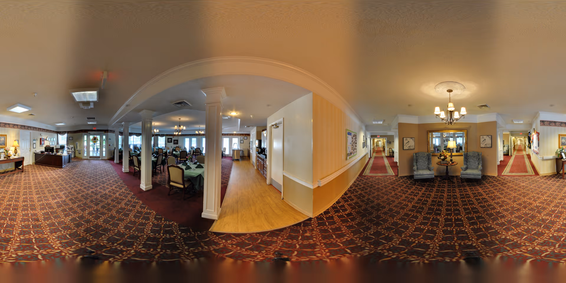 Panoramic interior showing a carpeted dining room and lounge area with tables, chairs, chandeliers, and adjoining hallways.