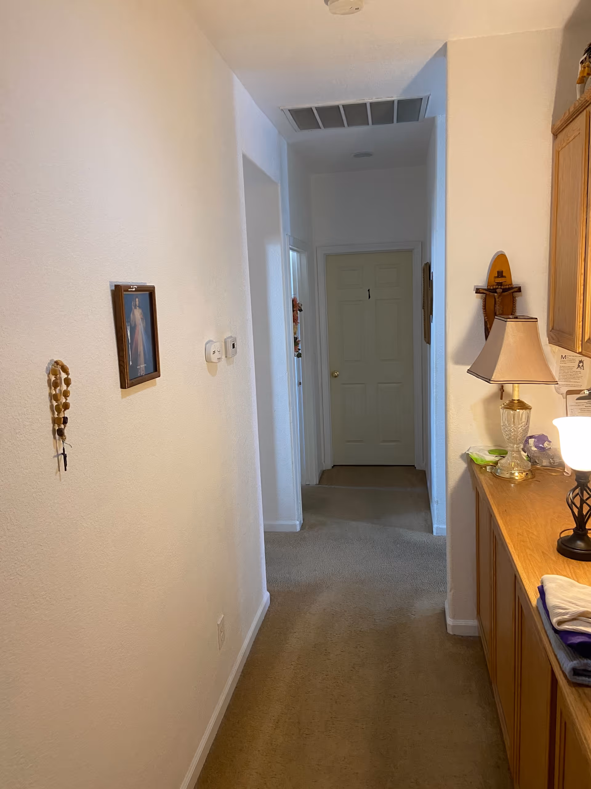Narrow interior hallway with carpet leading to a closed door, wall decor on the left and a lamp and cabinets on the right.
