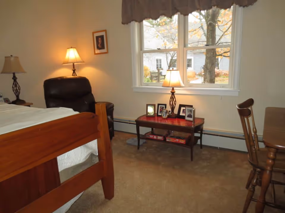 A cozy bedroom with a wooden bed frame, a dark brown recliner chair, two table lamps on either side, a small wooden table with framed photos and board games underneath, a window with a view of trees and a building outside, and a wooden chair next to a table.
