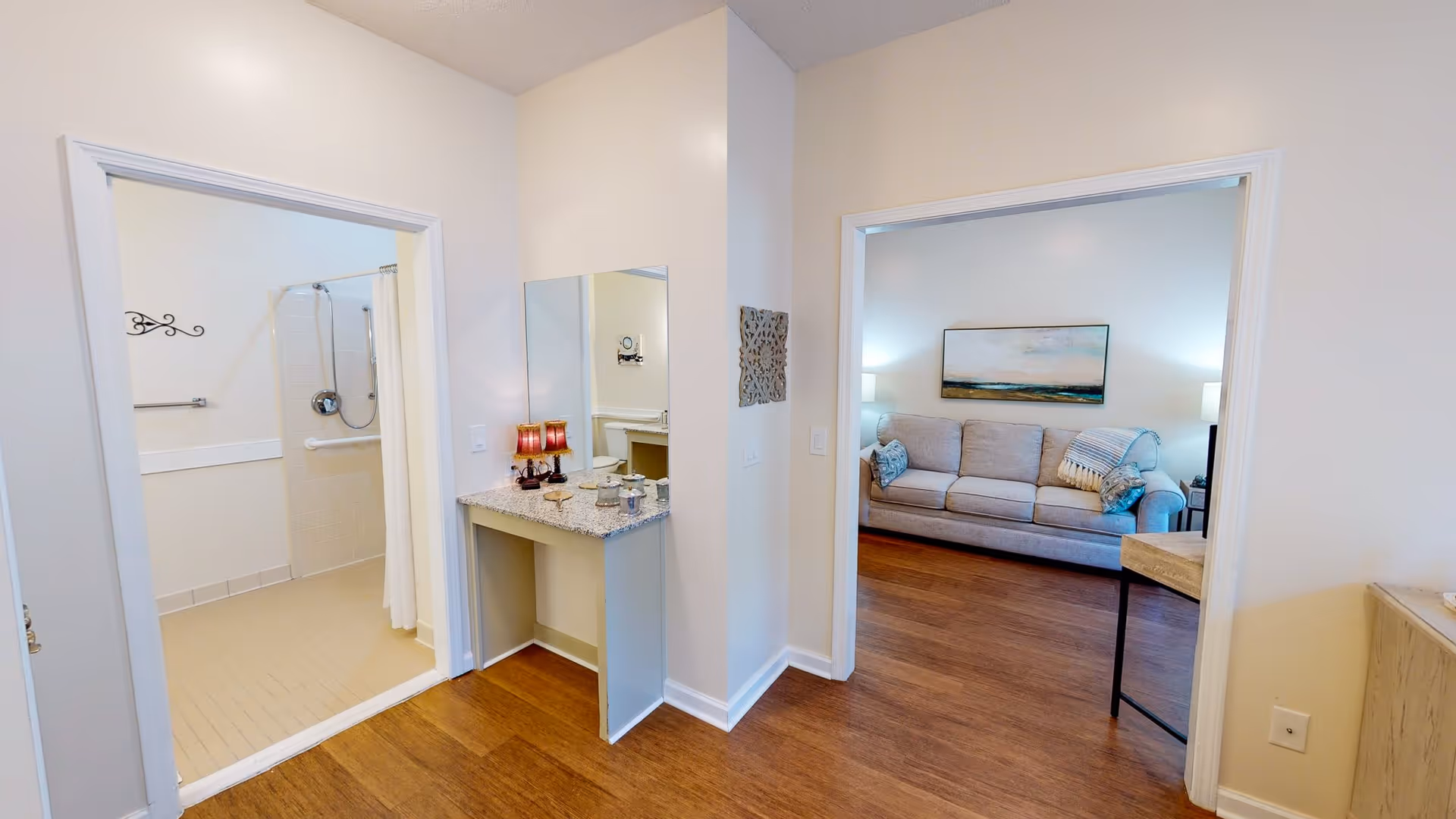 View of a hallway in a senior living facility showing an open bathroom with a walk-in shower on the left and a living room with a gray sofa, two side tables with lamps, and a landscape painting on the wall on the right. The hallway has a small vanity with a mirror and decorative items on the countertop.