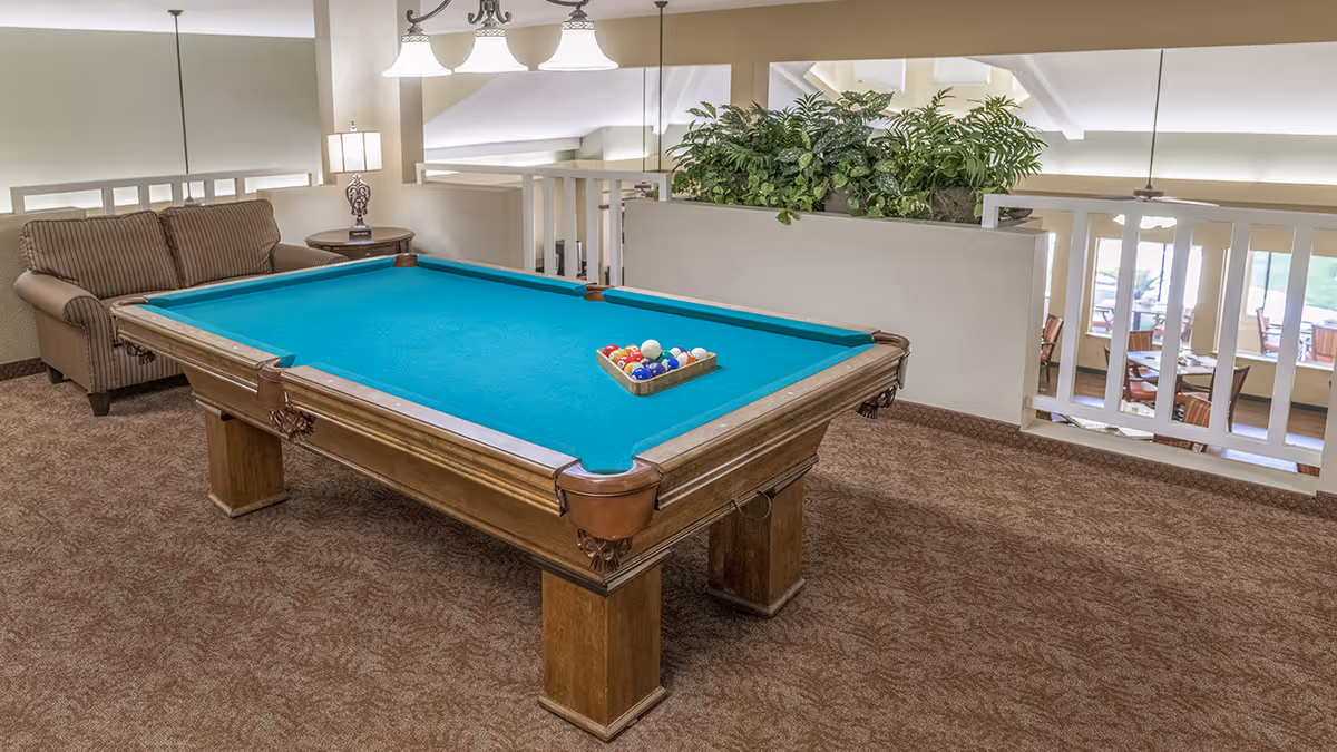 Billiards table with teal felt in a carpeted lounge area next to a sofa, side table, hanging lights, and a planter overlooking a dining space.
