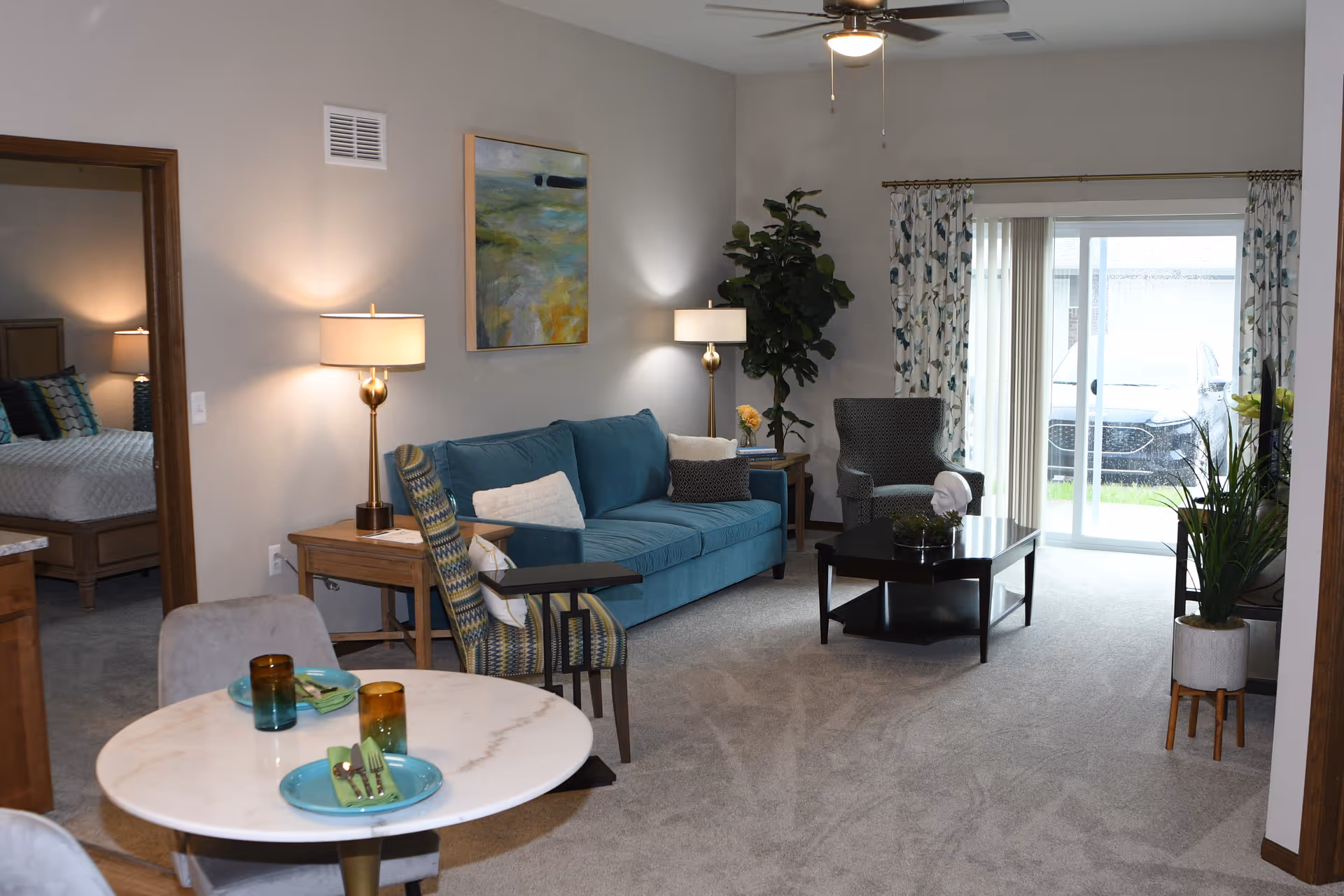 A cozy living room with a blue sofa, patterned armchair, coffee table, and side tables with lamps. There is a round dining table with two place settings in the foreground. A ceiling fan is mounted above, and a sliding glass door with floral curtains leads outside. A bedroom with a bed is visible through an open doorway on the left.