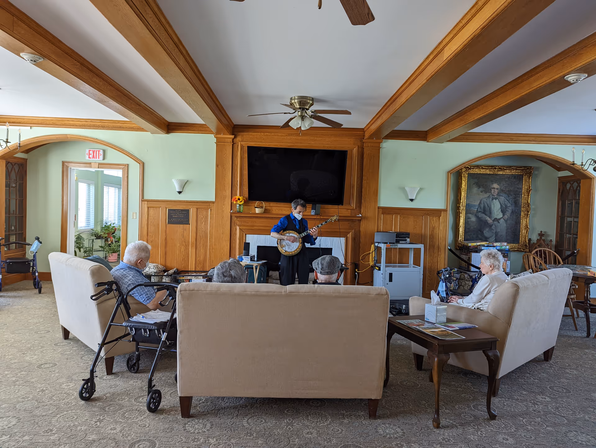 Several elderly residents sit on sofas in a living room while a musician plays a banjo near a fireplace and TV.