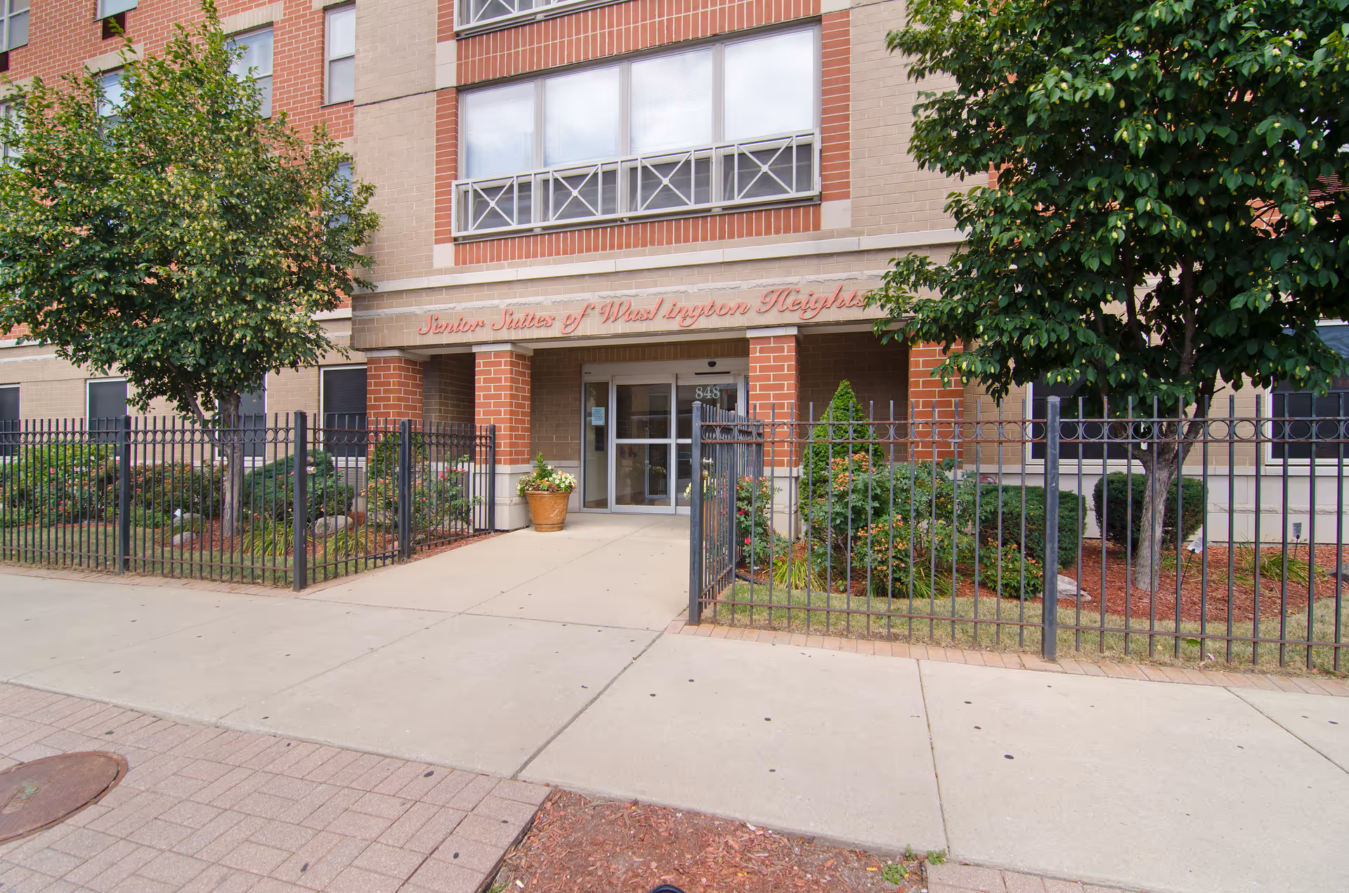 Entrance of a senior living facility named Senior Suites of Washington Heights, featuring a brick and beige exterior, a glass door, a black metal fence, and landscaped greenery with trees and shrubs.