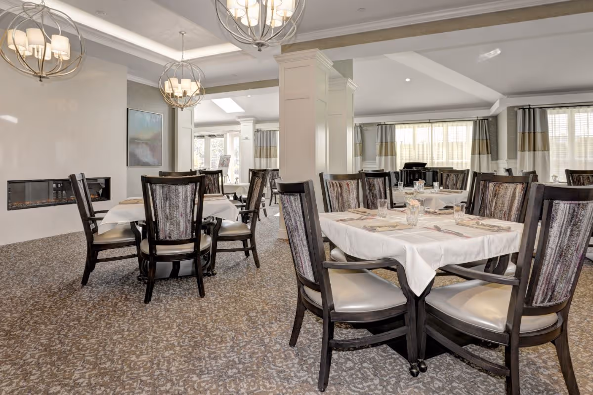 A well-lit dining room with multiple tables covered with white tablecloths, each set with glasses, napkins, and silverware. The room features patterned carpet, upholstered chairs with wooden frames, modern spherical chandeliers, large windows with curtains, and a built-in wall fireplace.