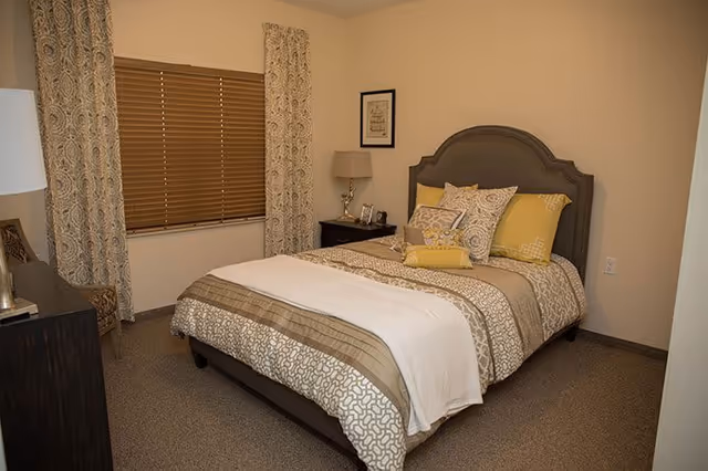 A neatly made bed with patterned linens and accent pillows in a cozy bedroom with a nightstand, lamp, and window with blinds and curtains.