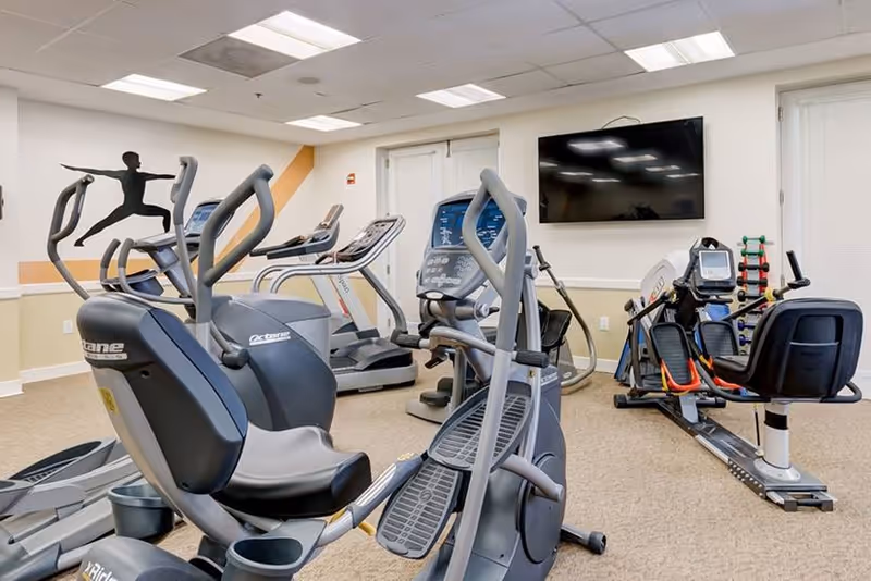 Indoor fitness room with exercise equipment including stationary bikes, treadmills, and a rowing machine. A wall-mounted flat screen TV is visible, along with a rack of small dumbbells. The room has beige carpet, white walls with a beige and orange accent, and a ceiling with fluorescent lighting.