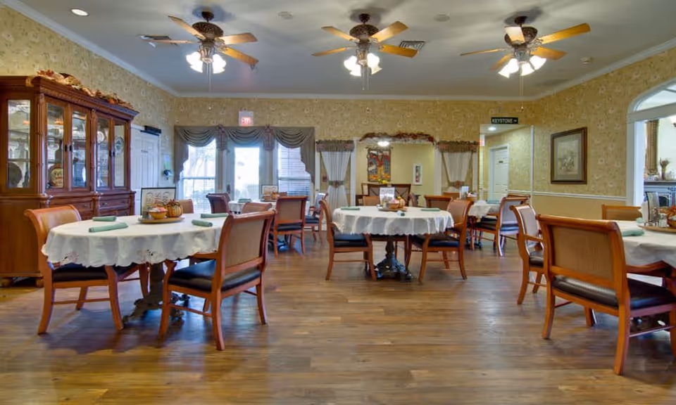 A senior living facility dining room with round tables covered in white tablecloths, each set with green napkins and decorative centerpieces. Wooden chairs with cushioned seats surround the tables. The room features wooden flooring, floral wallpaper, ceiling fans with lights, and a wooden china cabinet on the left side. Large windows with curtains allow natural light to enter the space.