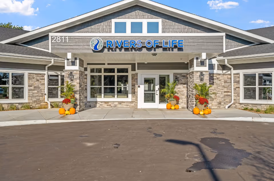 Front exterior view of the Rivers of Life Memory Care/Enhanced Assisted Living facility with a gray stone and siding facade, large windows, a white entrance door, and fall decorations including pumpkins and potted plants on either side of the entrance.