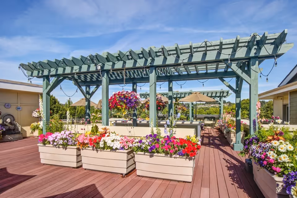 Outdoor patio area with wooden pergolas, hanging flower baskets, and large planters filled with colorful flowers. There are tables with umbrellas and chairs under the pergolas, set against a backdrop of trees and a clear blue sky.