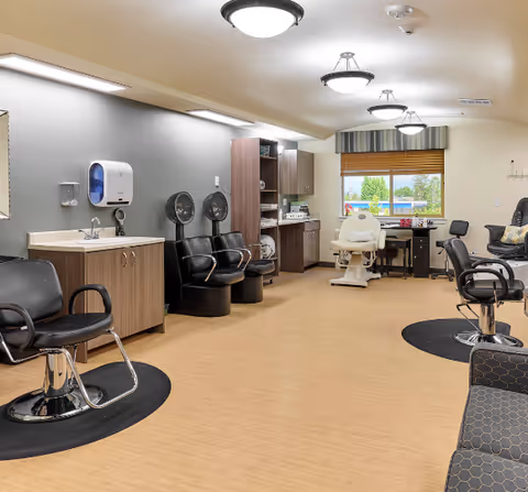 A spacious salon area in a senior living facility with multiple black salon chairs, hair drying stations, a sink with cabinets, and a manicure or pedicure chair near a window with a striped valance. The room has light wood flooring and neutral-colored walls with overhead lighting.