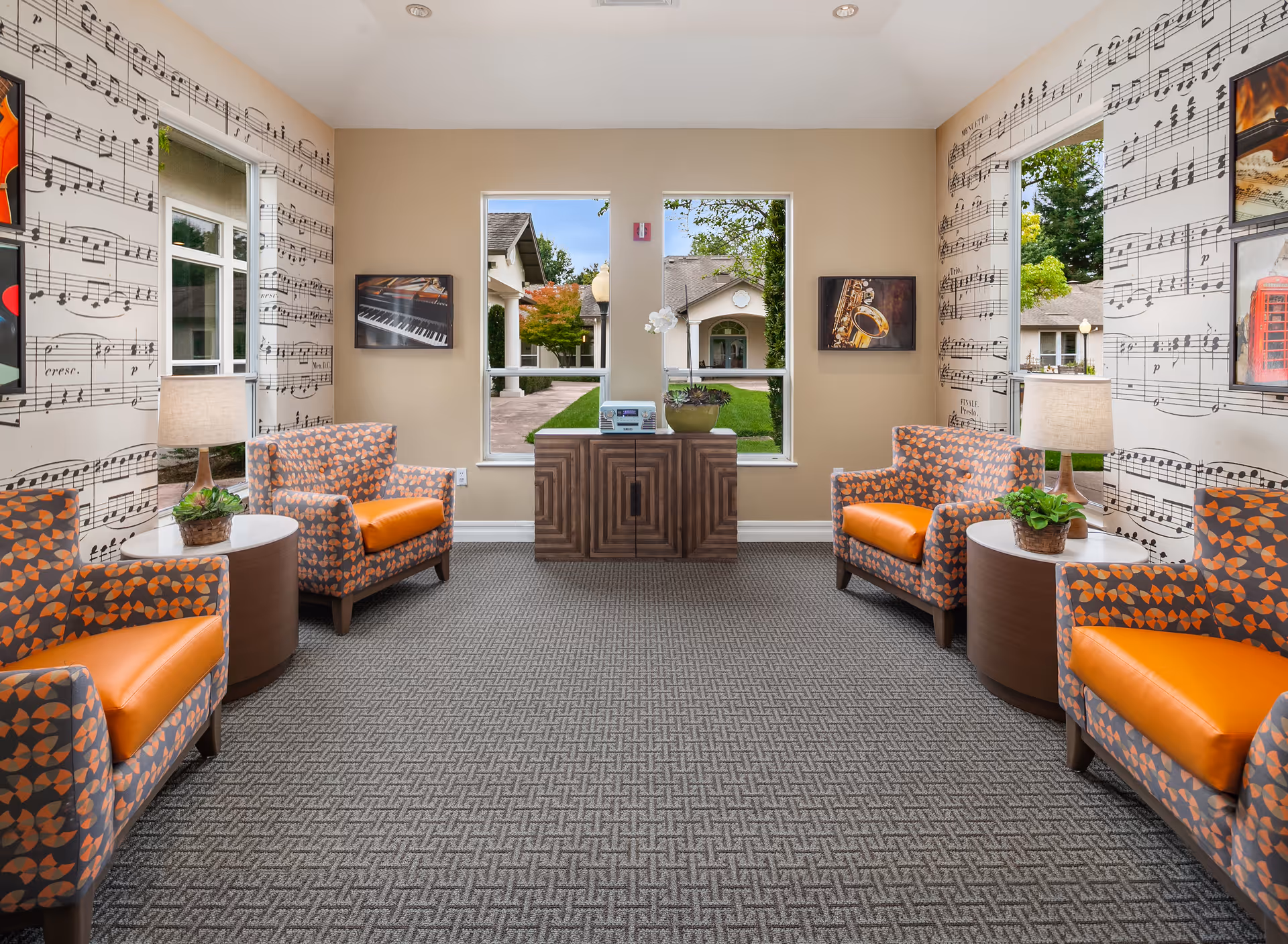 A bright and inviting sitting area with four patterned armchairs featuring orange cushions arranged around two round side tables with lamps and small plants. The walls are decorated with musical note wallpaper and framed pictures of musical instruments. Two large windows provide a view of a courtyard with greenery and buildings outside.