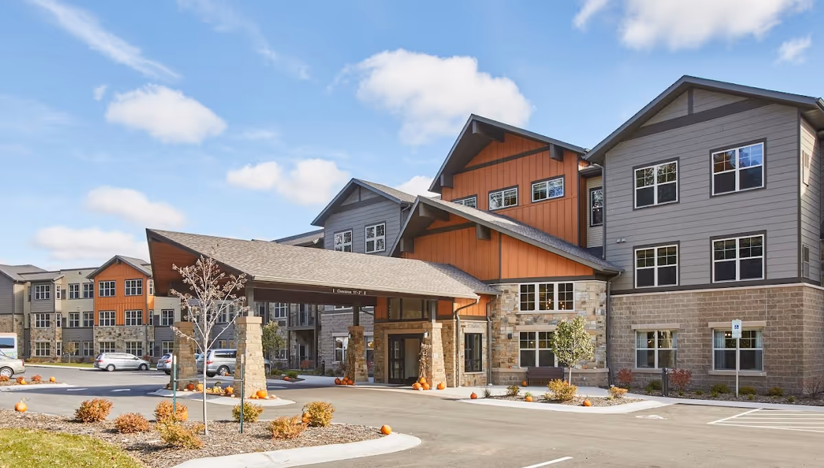 Exterior view of a senior living facility named Eagle Point Senior Living, featuring a multi-story building with a covered entrance, stone and wood siding, several windows, a parking lot with cars, and landscaping with small trees and bushes under a partly cloudy sky.