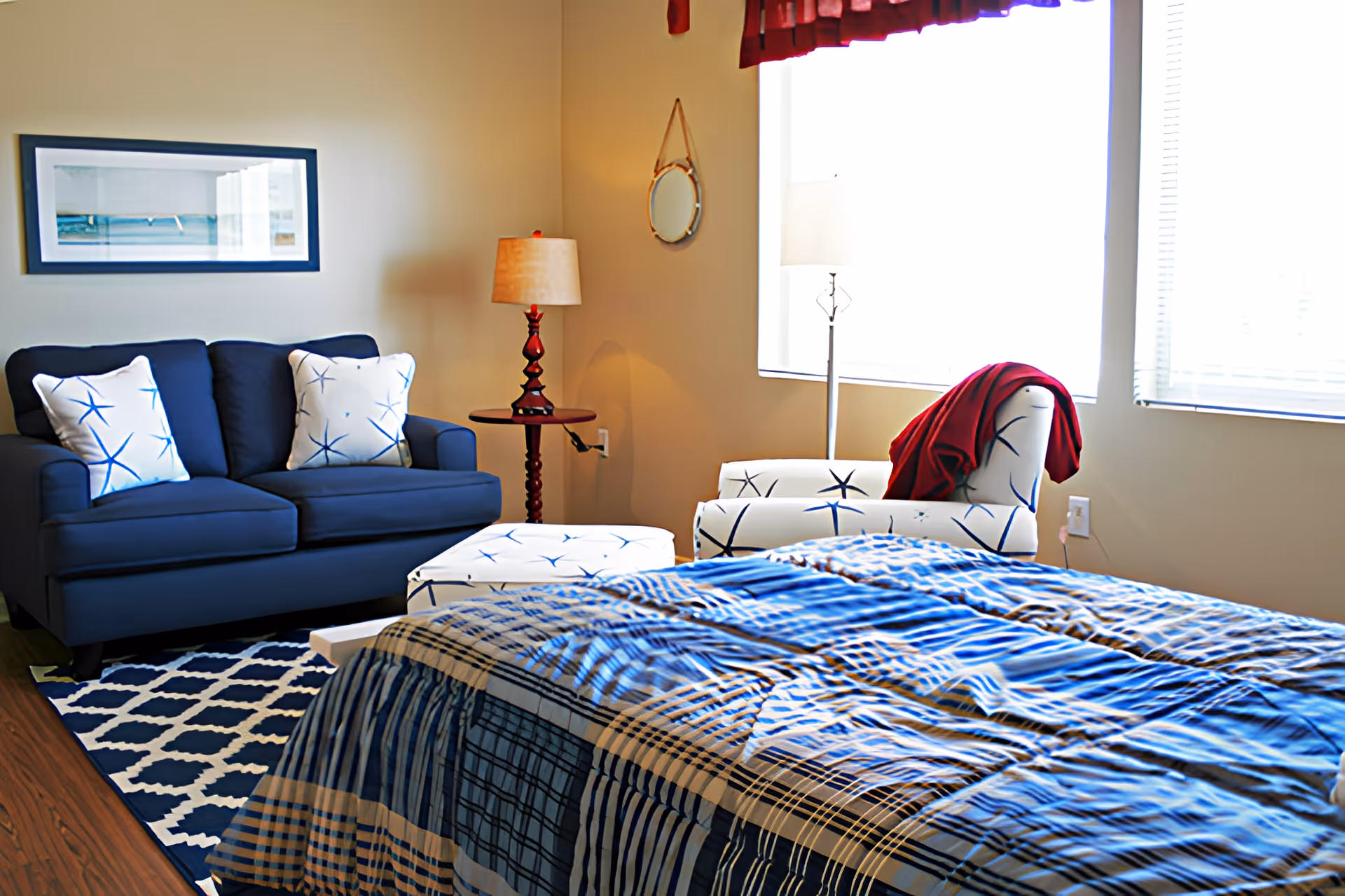 A bright bedroom featuring a bed with a blue patterned comforter in the foreground, a navy blue loveseat with two white starfish-patterned pillows, a matching ottoman, a white armchair with a red blanket draped over it, a wooden side table with a lamp, a small round mirror on the wall, and two large windows with blinds and a red valance.