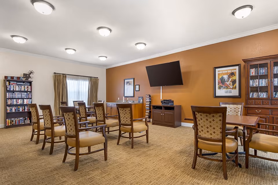 A well-lit common room with multiple wooden chairs arranged around tables, a large flat-screen TV mounted on an orange accent wall, bookshelves filled with DVDs and books, and framed artwork on the walls. The room has beige carpeting and several ceiling lights.