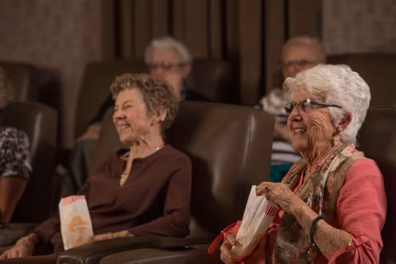 A group of elderly women sitting in comfortable theater-style seats, smiling and holding popcorn bags, enjoying a movie or presentation in a dimly lit room.