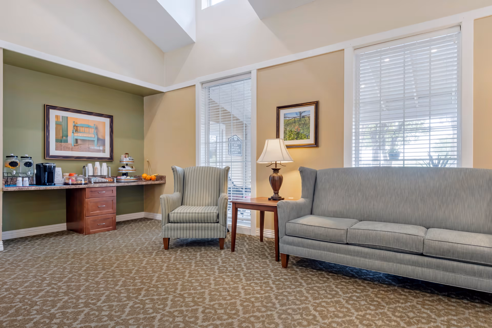 Well-lit communal living room with a sofa and armchair near a side table and lamp, and a countertop coffee/snack station against the wall.