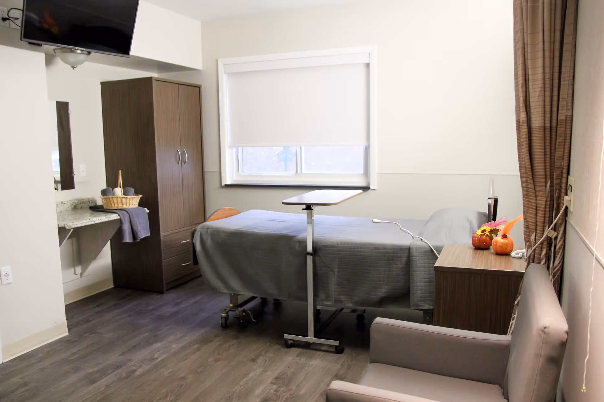 A simple, clean patient room with a hospital bed covered in a gray blanket, a small overbed table, a wooden wardrobe, a nightstand with small decorative pumpkins, a gray armchair, and a window with a white roller shade. The room has wood-look flooring and neutral-colored walls.